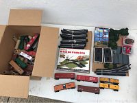 Top-down view showing box with multiple model freight cars and cardboard tubes, along with grouped curved track pieces, Absolute HO Scale Dual Crossing Gates box, bridges, and shrubbery pieces.