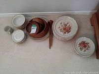 Stoneware dishes and wooden salad bowl set arranged on floor, showing variety of plates, bowls, cup, and wooden salad servers.