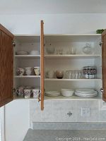 View of wooden cabinet open showing floral mugs and teacups with saucers on shelves along with plates and bowls on bottom shelf.