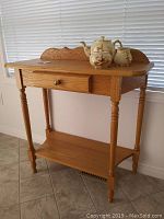 Oak single drawer table with turned legs, lower shelf, brass knob drawer, placed against window with blinds.