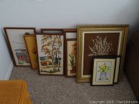 Nine framed embroidery pieces leaning against wall in various sizes and frame styles.