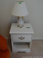 White wooden night table with one drawer, ornate metal handle, and open lower shelf with white ceramic lamp on top with gold accents and white pleated lampshade.
