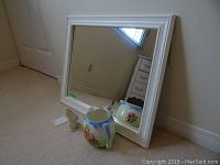 White framed square mirror resting against wall with ceramic pitcher and small bird figurine in front.