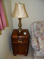 Wooden cabinet with one drawer and one door, dark finish, next to floral patterned sofa with brass look lamp on top