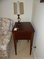 Photo of a wooden sewing machine cabinet with a false drawer front, medium brown finish, next to a floral upholstered couch, with a table lamp on top.
