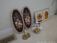 View of entire lot showing two framed embroidery pieces, two oval ceramic floral wall hangings, small anniversary clock with dome, and ceramic pitcher on doily on carpeted floor against wall.