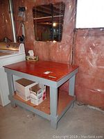 Wooden utility table with reddish wood top and gray painted legs and lower shelf. The table sits on a basement floor near a washing machine.