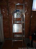 Two aluminium ladders standing vertically in a room with insulation walls: one taller 5 ft ladder and one shorter 2-step painter's ladder with platform and handle bar.