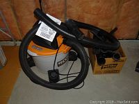 Photo of Ridgid shop vacuum model WD945 with hose loosely wrapped and some vacuum attachments nearby on floor.
