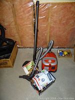 Wide shot of orange-red Miele canister vacuum with grey hose, metal wand, user manual, box of new vacuum bags and attachments on concrete floor