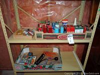 Two cardboard boxes on a shelf filled with assorted small handyman supplies including packaged screws, nails, wood filler, and a caulking gun.