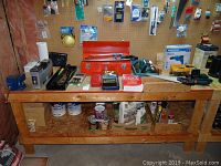 Wide shot of wooden workbench with red metal tool box, saws, socket set, and assorted small tools on and underneath, showing items included in the lot in a workshop setting.
