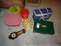 Lot overview showing all items together on the floor including the vintage Coleman cook stove, Thermos cooler bag, large thermometer, wooden barometer, small orange waste can, and pink floral cushioned stool.