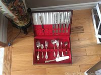 Wooden box open with red lined compartments holding 5 forks and 5 soup spoons with ornate floral handles, additional cutlery pieces under lid.