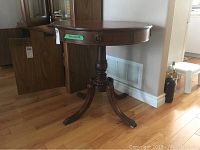 Round wooden pedestal table with reddish-brown leather top, decorative brass hardware on edge, four outward curving legs on a turned pedestal base shown from angled side.