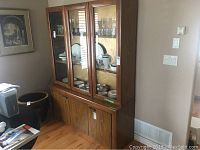 Wood and glass china cabinet, showing the side angle with the left wall and part of surrounding furniture.