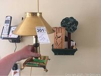 Handheld brass tri-light lamp with conical shade, label attached, cordless phone and wall-mounted key holder visible in background