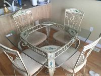 Front view of the glass top table and four chairs arranged around it showing glass tabletop, decorative metal base, and chair details.
