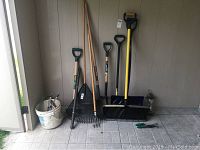 Photo showing assortment of garden tools including shovels, rakes, edger, bucket with accessories.
