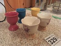 Three egg cups placed on carpet; two cream-colored with floral and basket-weave design, one pink smooth ceramic cup.