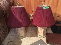 Two tri-light lamps with maroon pleated shades and Asian motif ceramic bases shown side by side against a wood-paneled wall and carpeted floor.