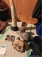 Lot contents showing tall carved ivory-colored sculpture, carved sheepdog, decorative tile, small ornate frame, and Honeysuckle Cottage figurine on carpeted floor.
