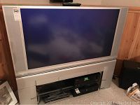 Front view of Hitachi 50 inch CRT TV including integrated stand and shelving below the screen. The TV is silver/gray with large side speakers.