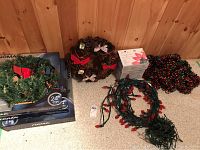 Photo showing three Noma pre-strung pine wreaths with red bows on boxes, one pinecone wreath with small decorations and bow, and two coiled Christmas light strings and red-green garland on floor.