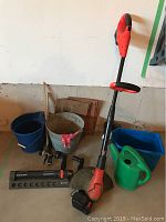 Overview of battery weed trimmer standing upright with various buckets, watering can, sprinkler, hose nozzles, and gardening tools around it.