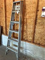 Full height view of a 6-foot aluminum step ladder leaning against wooden wall, showing multiple steps and wear marks.