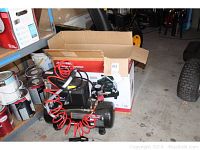 Husky air compressor shown on concrete floor near shelving with paint cans, showing red coiled hose and control gauges.