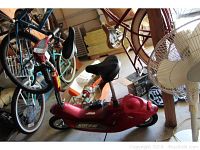 Full view of the red Scoot N Go electric scooter with a bicycle-style seat, handlebars, and small wheels, surrounded by bicycles in a garage environment.