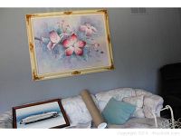 Photo showing original oil painting with pink and purple flowers in a decorative frame above a couch, and framed poster leaning against the couch.