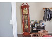 Full view of tall Ridgeway grandfather clock against wall next to bookshelf and coats.