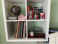 White shelving unit with four cubical compartments holding various items including books, vinyl records, a turntable, a pink elephant bank, a black globe, and a decorative phrase sign.