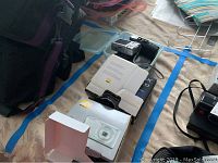 Overview of the lot showing multiple boxes and items including the Nikon Coolpix S3700 packaging, Kodak EasyShare camera, and other items on a bed.