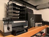 Photo showing HP Office Jet Pro K5400 printer with stacked paper organizer, Audiofile HT2000 home theater speakers including subwoofer and satellite speaker, black electronic device (likely Direct TV box), and a monitor on wood shelf.