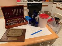 Photo of wooden shelf, metal wire rack, kitchenware including blue glasses, plastic travel cups, silver plate flatware in wooden box, and John Deere plaque.