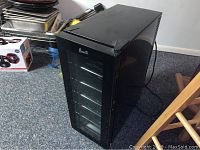 Front and angled view of black Avanti wine chiller fridge with glass door and metal wire racks inside, placed on carpeted floor.