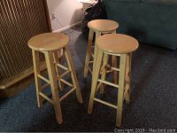 Photo showing all three wooden stools together from a slight angle, placed on carpet next to furniture and a lamp.