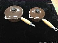Top and side view of two brown enamel Le Creuset sauce pans with wooden handles and matching lids on black background.