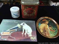Photo showing all four items in the lot arranged on a black cloth - red tobacco tin, white tea tin, Coca Cola oval tray with woman image, RCA reproduction tin sign showing His Master's Voice dog and gramophone.
