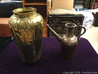 Brass vase and copper watering can side by side on a purple cloth with furniture in background.