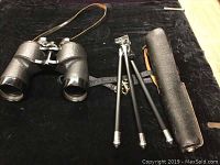 Full view of the binoculars, tripod, and carrying case arranged on a black cloth background.