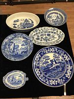 Overview of seven blue and white transferware pieces on black surface showing various patterns and shapes.