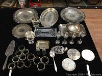 Photo showing the complete set of vintage silver plated serving trays, shakers, napkin rings, coasters, small vases, and serving utensils arranged on a black surface.