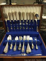 Top view showing knives and serving spoons secured in slots within hinged wooden chest with blue velvet lining.