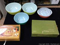 Two yellow mixing bowls and one green mixing bowl placed on black surface alongside a pink covered casserole, a butterfly gold covered casserole in original box, and an oval avocado divided serving dish with cover in original box.