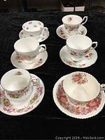 Photo showing all six vintage porcelain cups and saucers arranged on black fabric, each with distinct floral patterns and sizes.