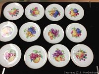 Set of 12 round fruit plates laid out on a black surface, showing colorful fruit illustrations and gold trim.
